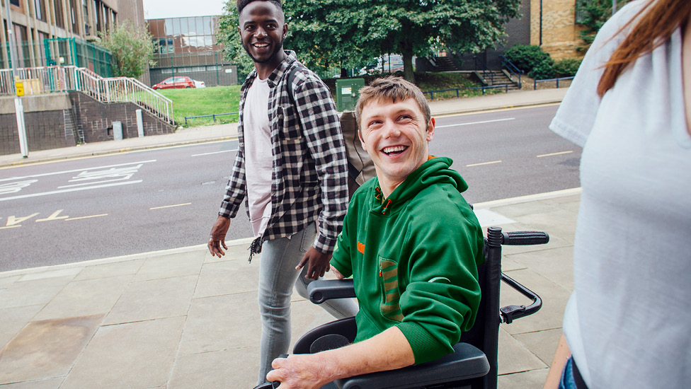 Young man in a wheelchair laughing with friends on a campus sidewalk.