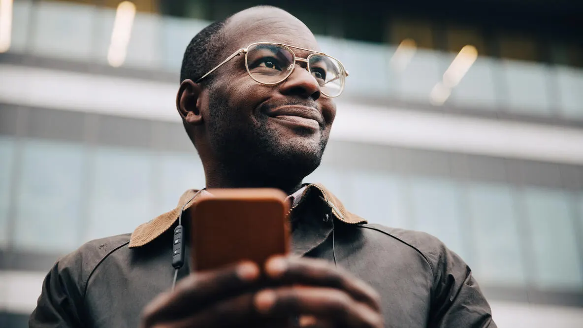 Low angle view of smiling businessman using smart phone looking away while standing in front of a building in city.