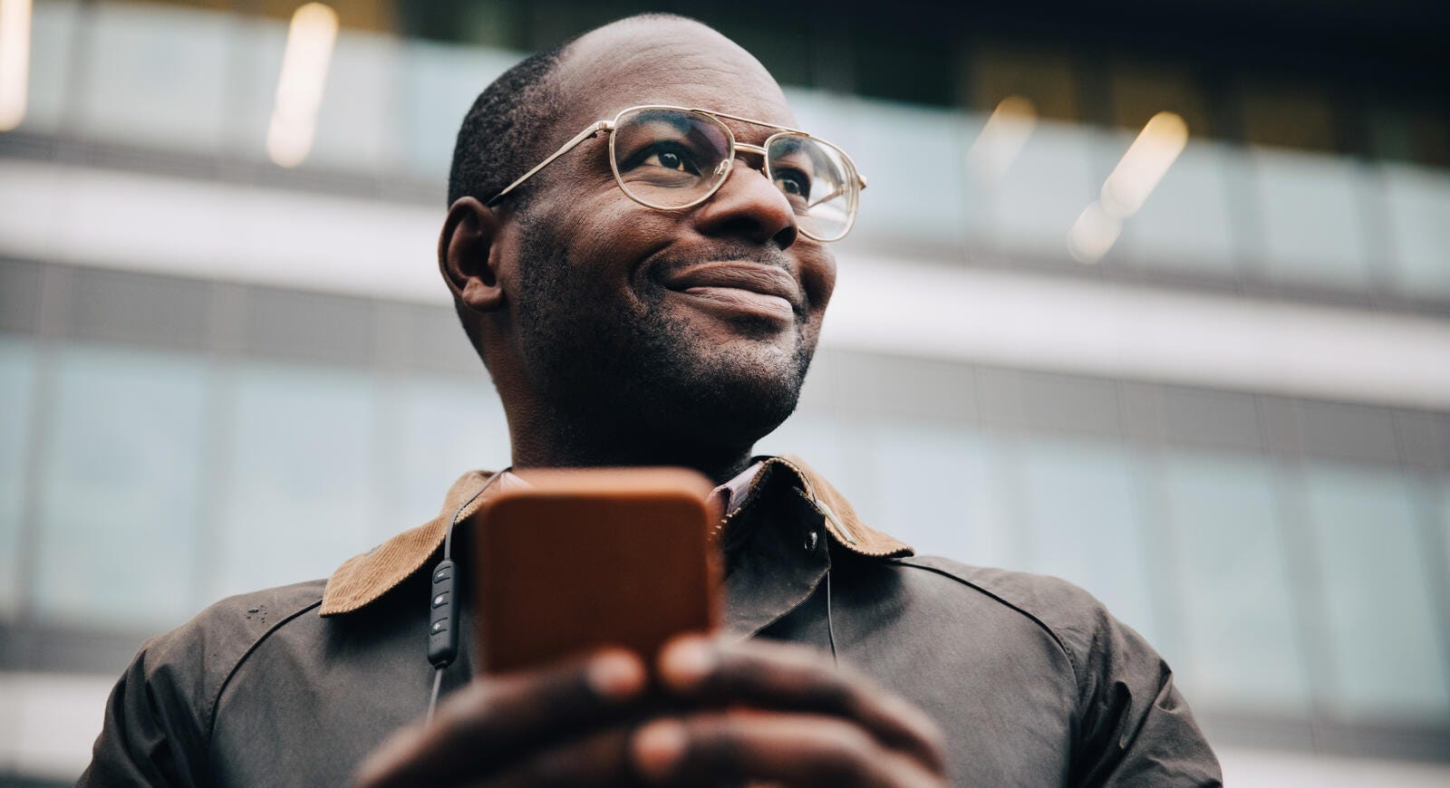 Low angle view of smiling businessman using smart phone looking away while standing in front of a building in city.
