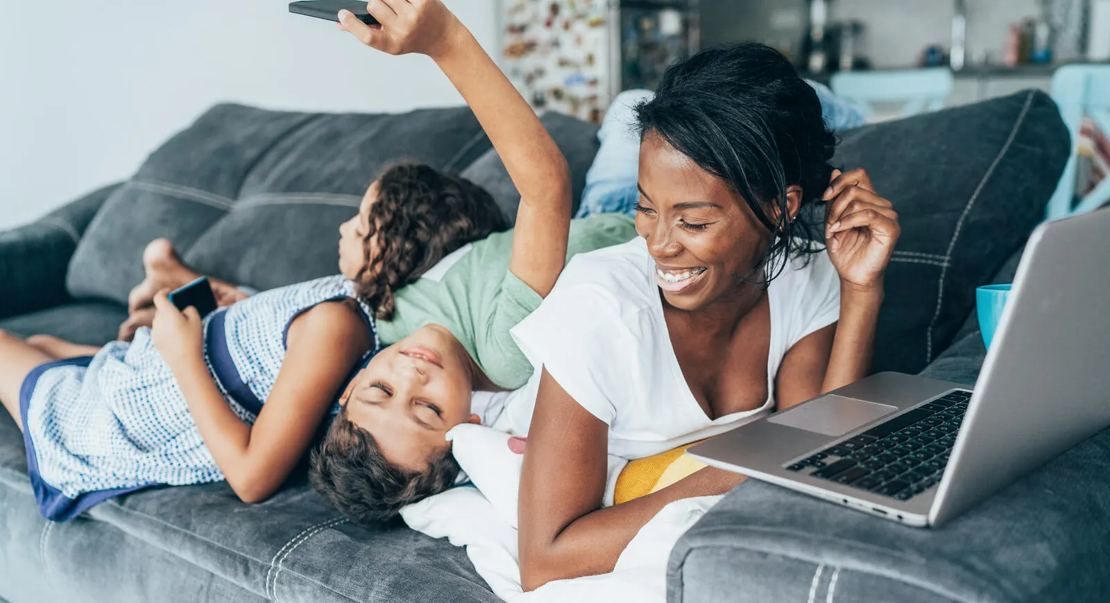 A mom laughing on the couch with her two children using smart phones and a laptop.