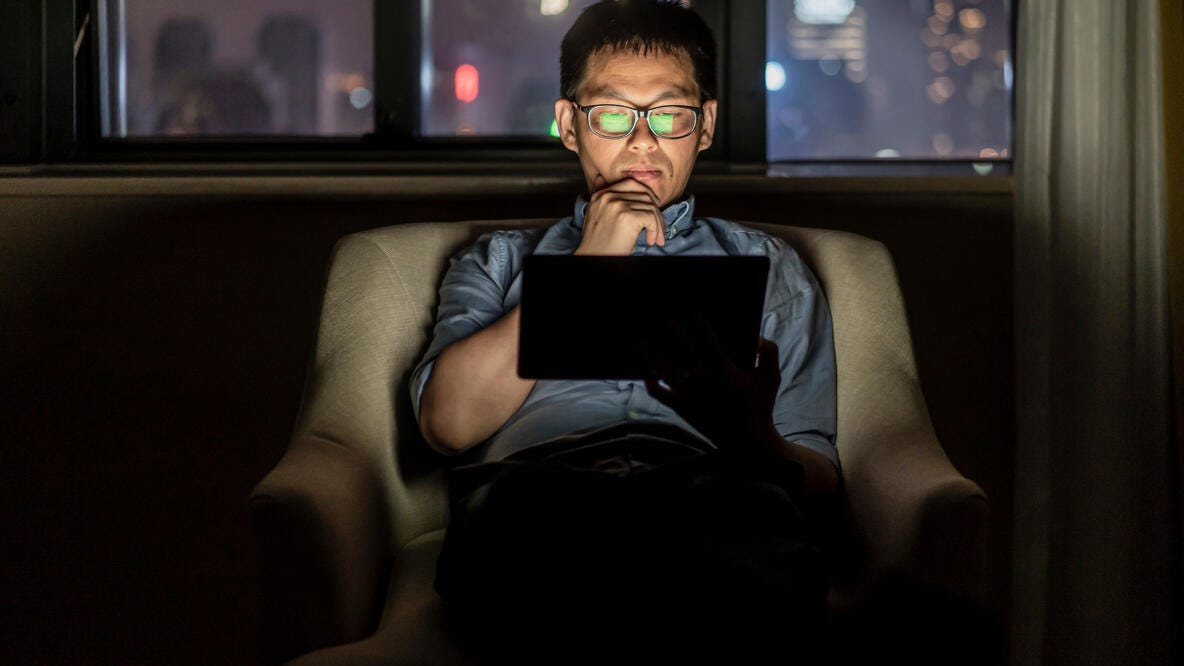 A man with glasses watching a tablet as he sits in the dark.