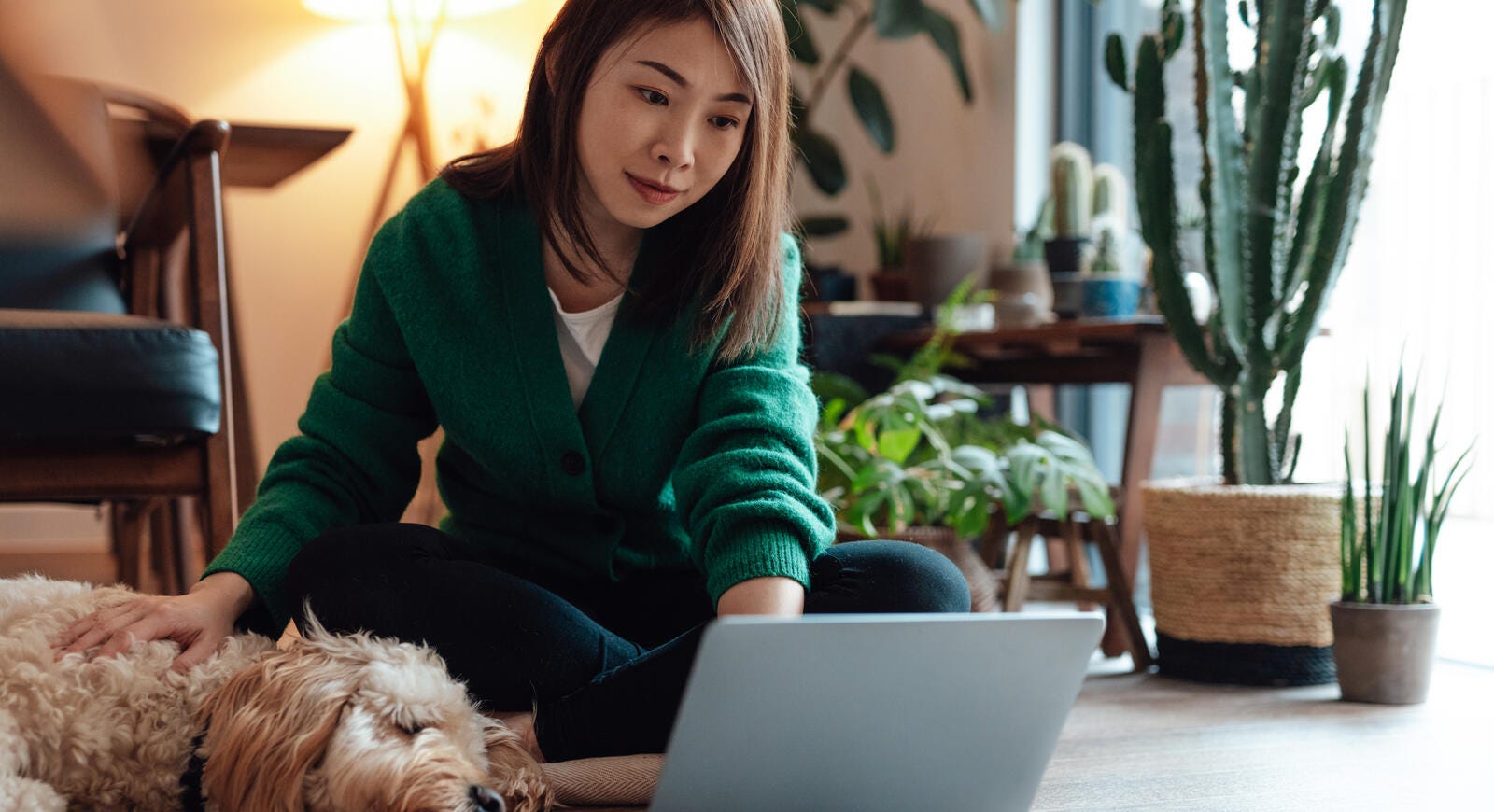 A woman sitting on the floor, petting her dog while using her computer.
