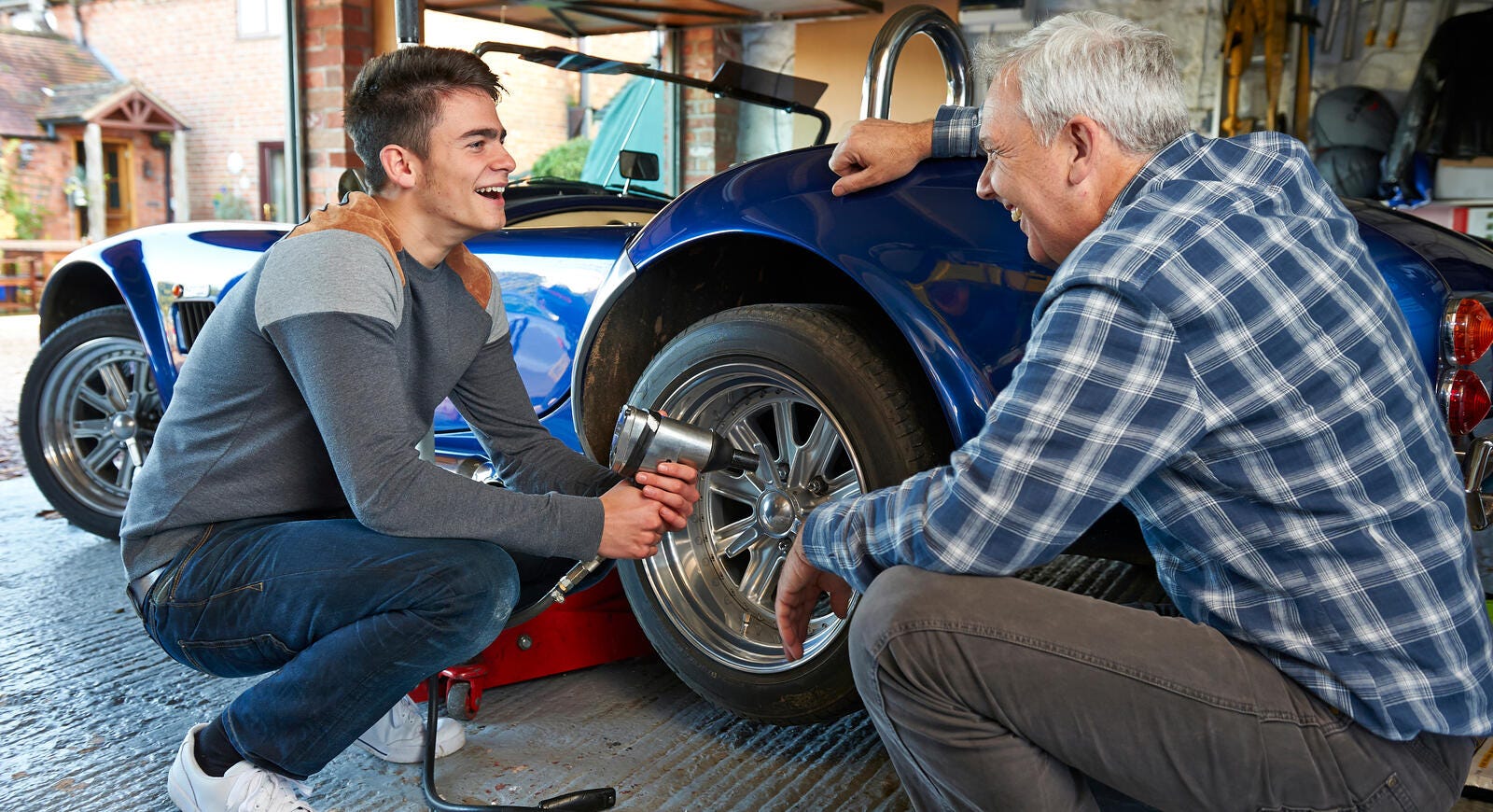 Father and teenage son laughing together while they change the tire on a blue classic Shelby Cobra.