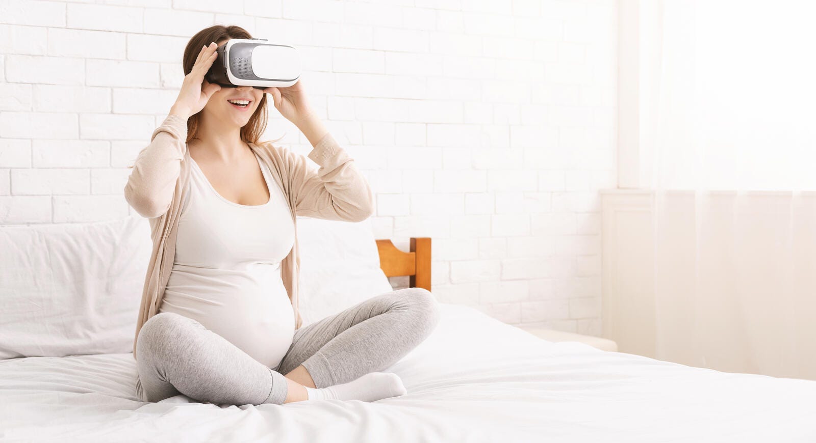 A pregnant woman sitting on a bed and smiling while wearing a virtual reality headset.