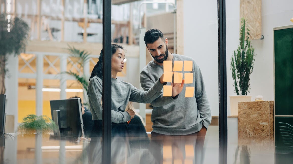 Two coworkers discussing and idea with sticky notes on a glass office wall.