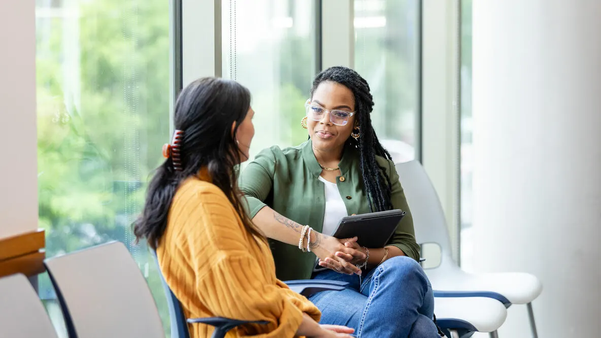 Two women having a conversation sitting in front of large windows.