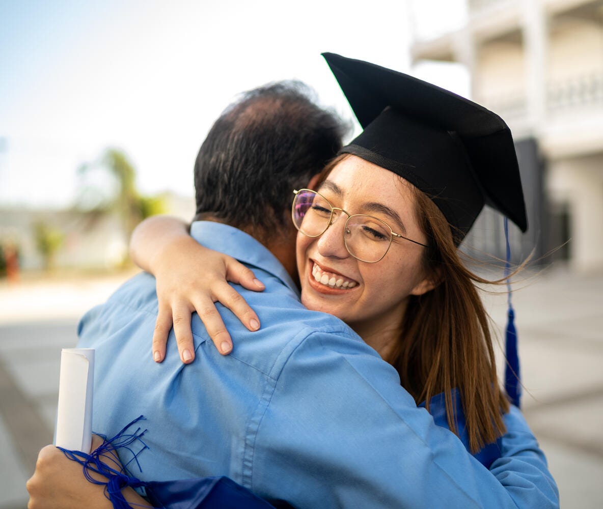 A girl wearing her hat and holding her diploma hugging her father after her graduation.