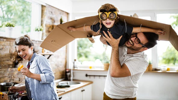 A child being held in the air by his father. The child is wearing pilot's goggles and has cardboard wings