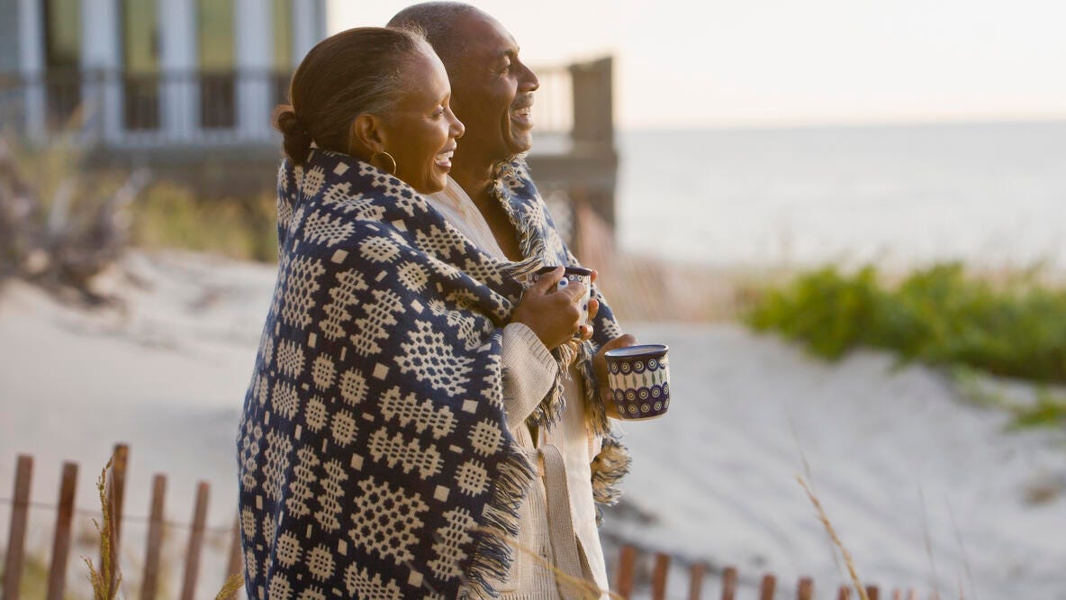 A couple standing together on a beach wrapped in a blanket, holding mugs and enjoying the ocean view.