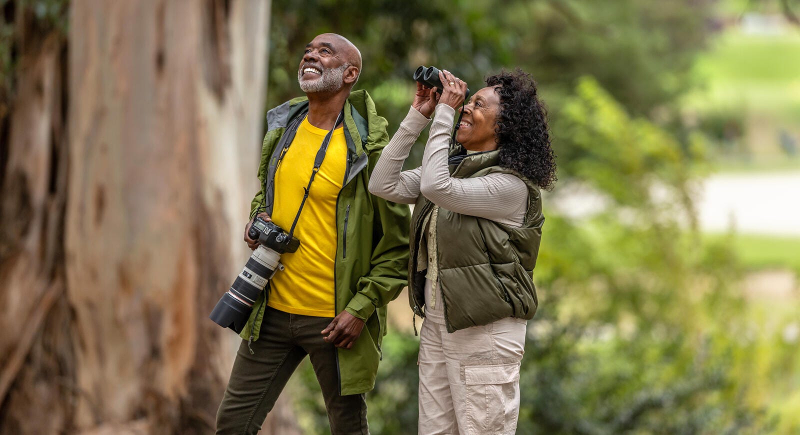 A retired couple bird watching in the woods with binoculars and a professional looking camera.