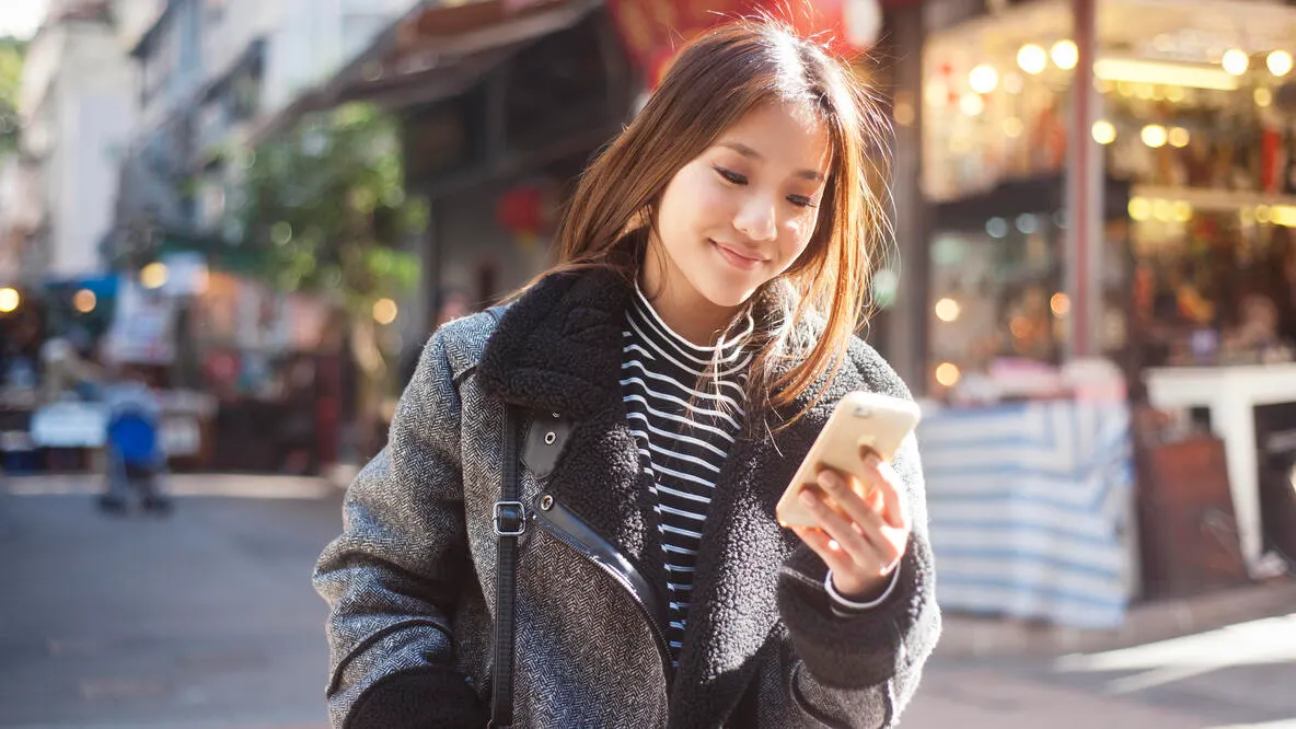 A smiling young woman using her phone while walking down the street.