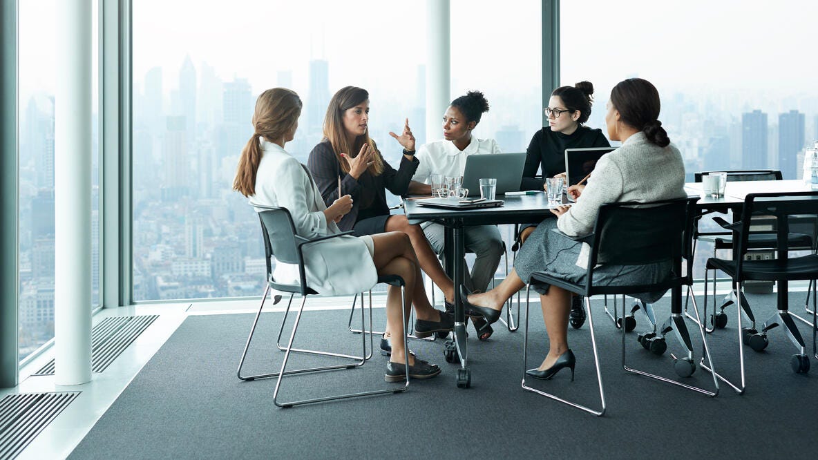 Five women in a conference room with floor to ceiling windows overlooking a city.