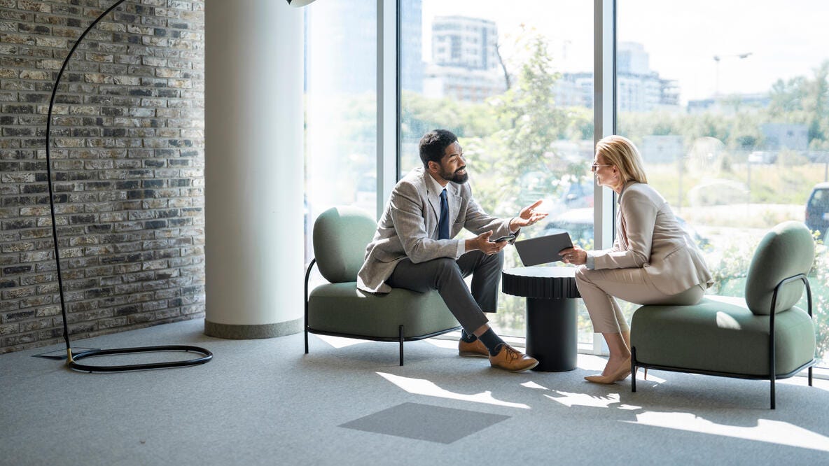 Two businesspeople are sitting in armchairs in a modern office lobby, discussing work and using a digital tablet.