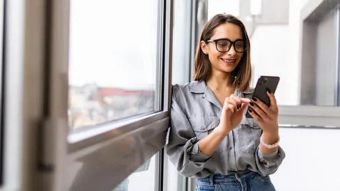 A woman smiling while using her cell phone in front of large windows. 