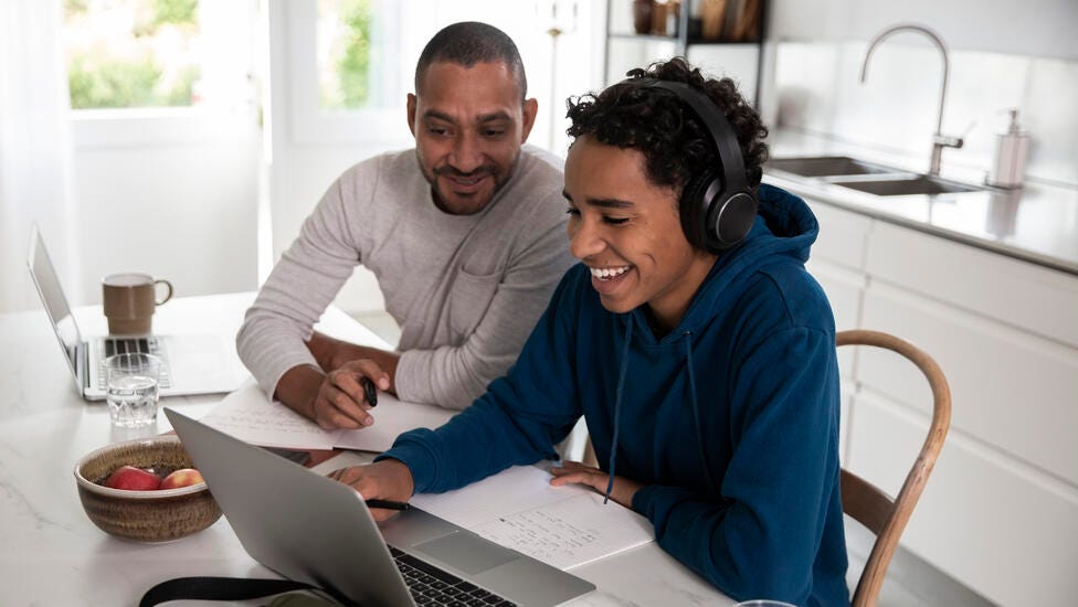 Muchacho joven utilizando una laptop junto a un adulto que hace anotaciones