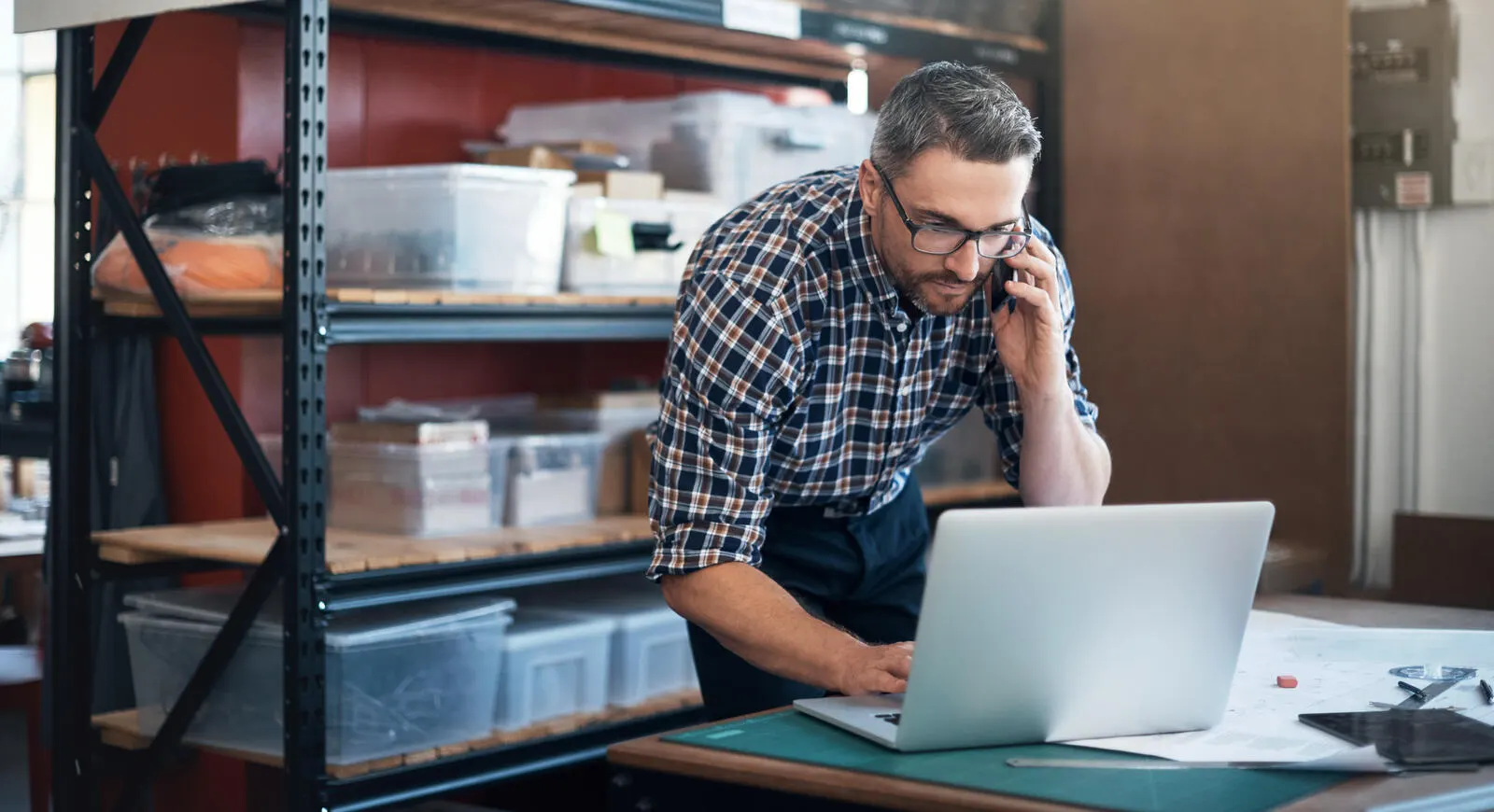 A man working on his laptop and cell phone in his workshop.