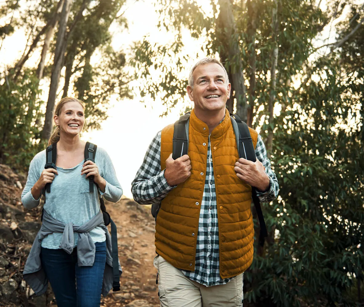 A happy couple hiking through the woods wearing backpacks.