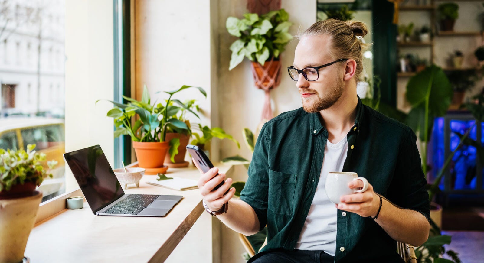 Man in a coffee shop looking at his phone with his laptop open in front of him