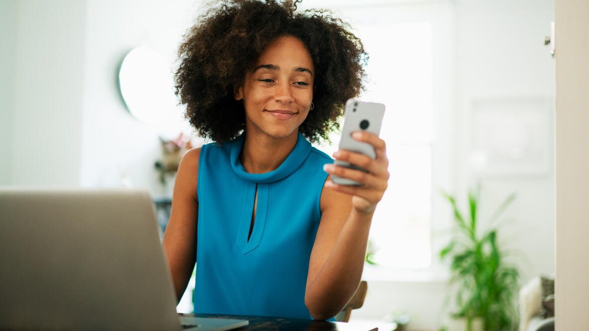 A young woman smiling while using her smartphone in a home office.