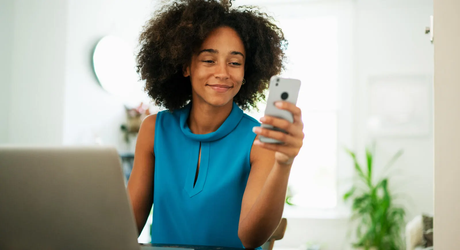 A happy young woman looking at her phone while also using her laptop.