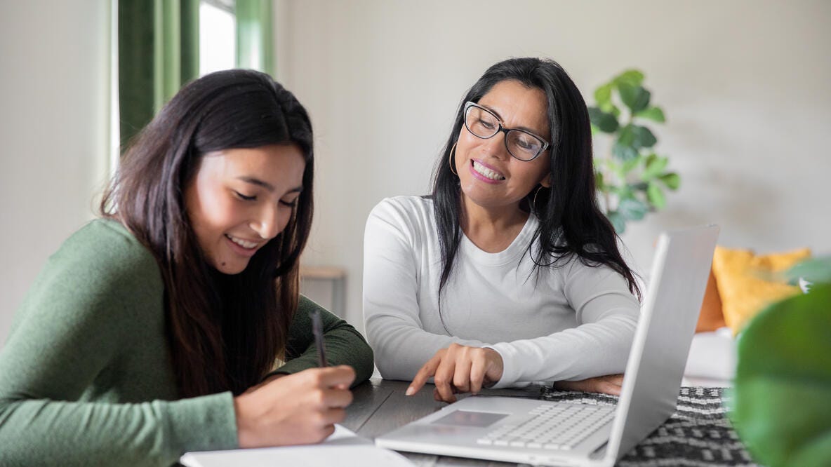 Muchacha joven utilizando una laptop y haciendo anotaciones junto a su madre que la asiste.