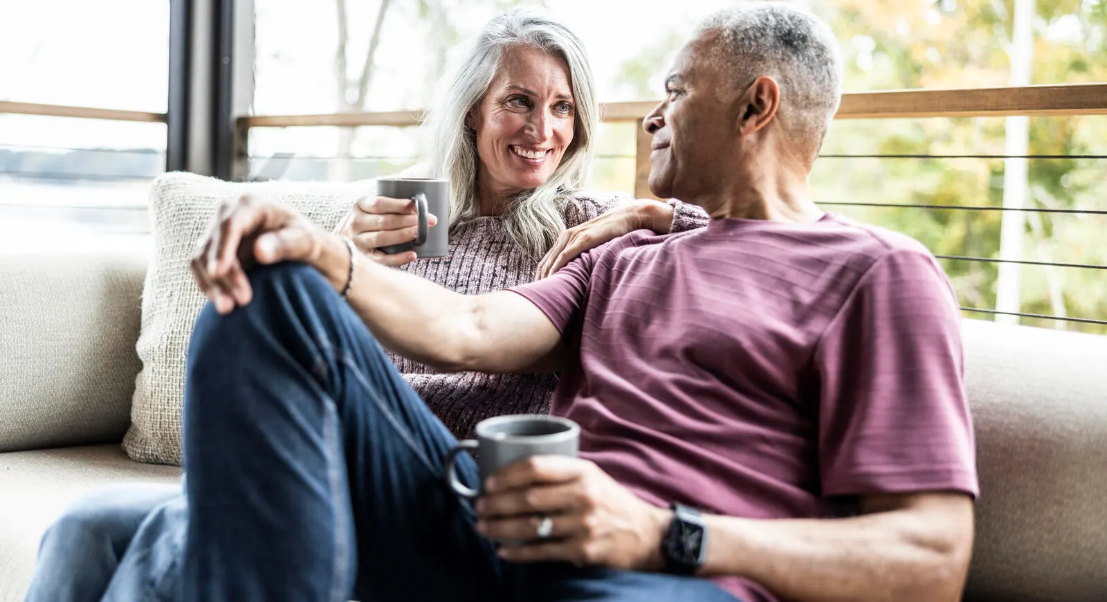 A couple sitting on the couch drinking coffee and smiling at each other. 