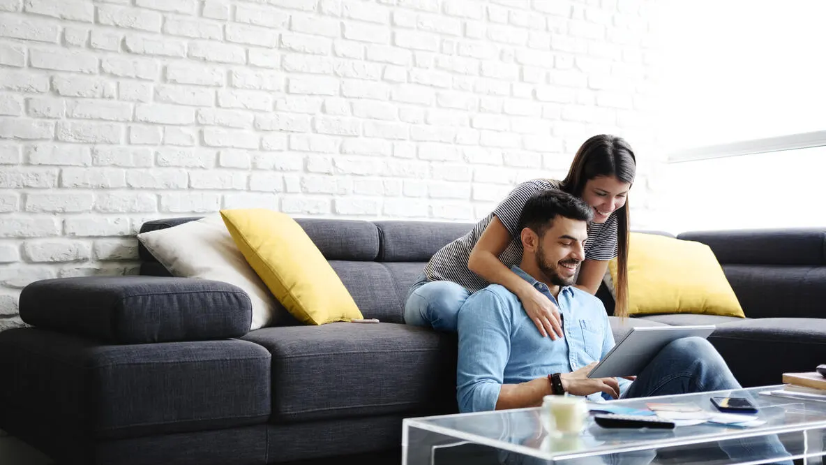 A smiling couple sitting on a couch reviewing finances on a tablet.